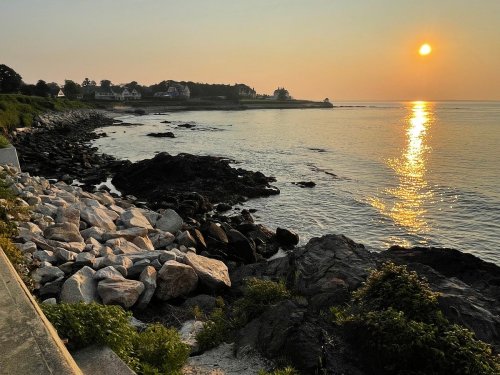 Sunset over a rocky shoreline, with calm waters reflecting the golden light and houses in the background.