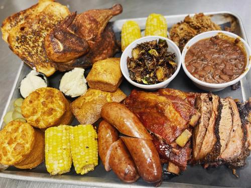 A metal tray filled with various meats, cornbread, corn on the cob, and sides like beans and Brussels sprouts.