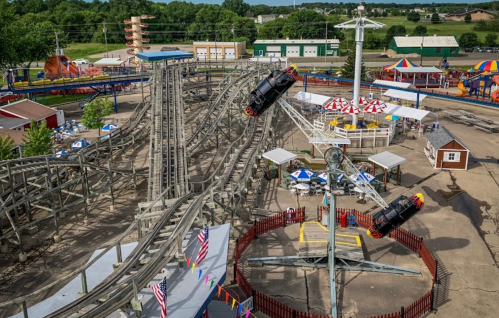 Aerial view of a theme park featuring a wooden roller coaster, rides, and colorful tents in a green landscape.