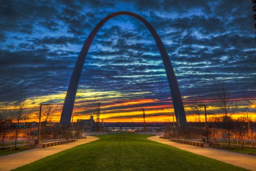 A stunning sunset behind the Gateway Arch, with vibrant clouds and a serene park pathway in the foreground.