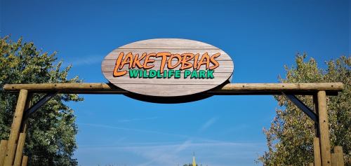 Sign for Lake Tobias Wildlife Park against a clear blue sky, framed by trees.