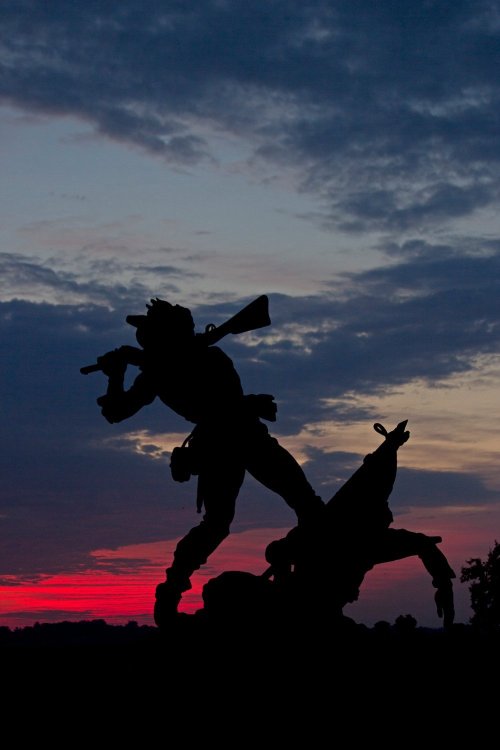 Silhouette of a soldier with a rifle against a colorful sunset sky, highlighting dramatic clouds and landscape.