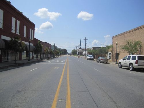 A wide, empty street lined with buildings and trees under a clear blue sky with a few clouds.
