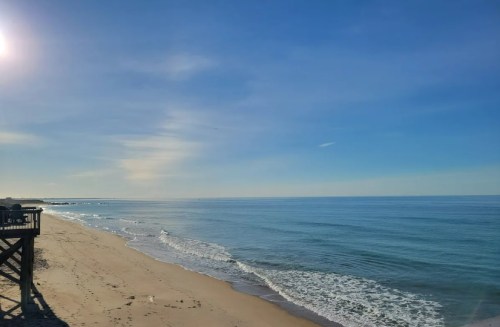 A serene beach scene with gentle waves, clear blue skies, and a distant pier under bright sunlight.