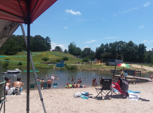 A sunny beach scene with people swimming and relaxing by a lake, surrounded by green hills and colorful umbrellas.