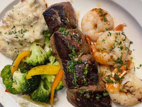 A plate featuring grilled steak, shrimp, mashed potatoes, and sautéed broccoli with bell peppers, garnished with herbs.