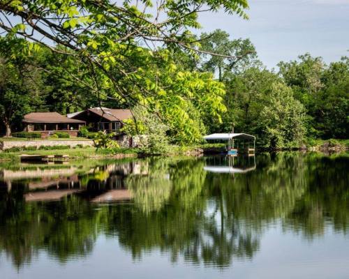 A serene lake reflects a house and trees, with a dock and boat lift visible on the water's edge.