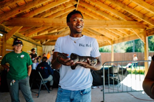 A smiling person holds a snake in a wooden shelter, with others in the background engaged in activities.