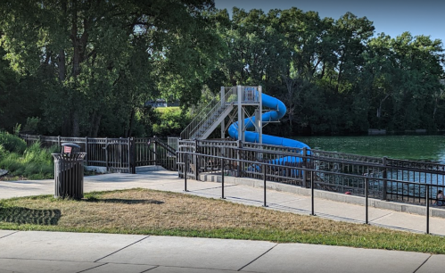 A blue water slide next to a river, surrounded by trees and a paved walkway with a trash can nearby.