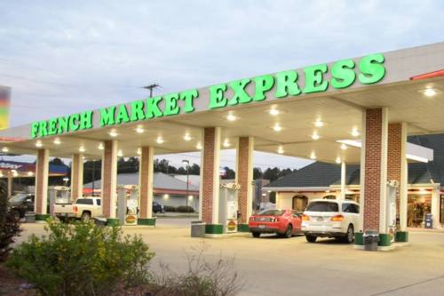 Gas station with a bright green sign reading "French Market Express" and cars parked in front. Evening setting.