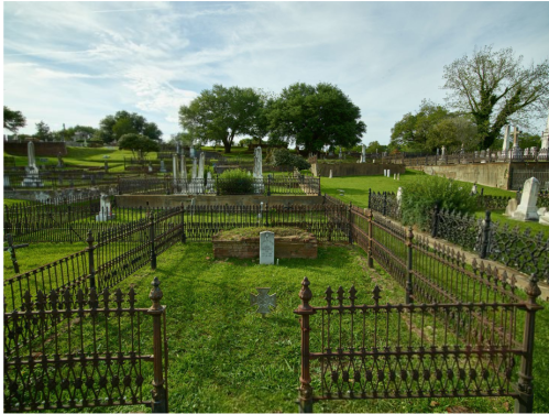 A peaceful cemetery with green grass, ornate iron fences, and gravestones under a blue sky with scattered clouds.