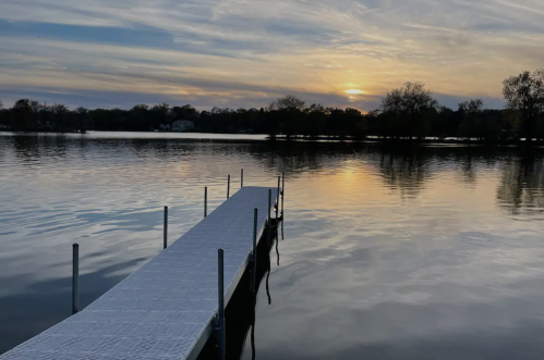 A serene lake at sunset, with a wooden dock extending into the calm water and trees silhouetted against the sky.