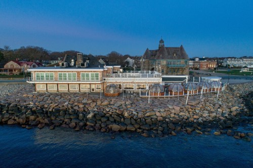 A waterfront restaurant on a rocky shore, with a historic building in the background and a clear blue sky.