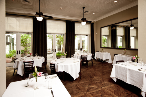 Elegant restaurant interior with white tablecloths, wooden chairs, and large windows letting in natural light.