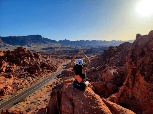 A person sits on a rocky outcrop, making a heart shape with their hands, overlooking a desert landscape at sunset.