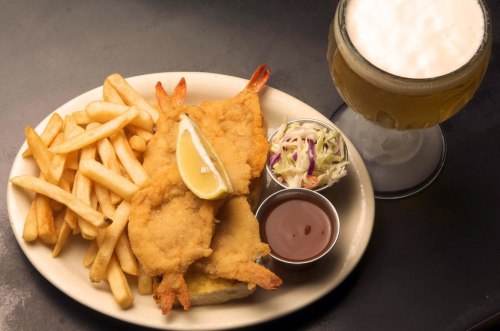A plate of fried shrimp with a lemon wedge, fries, coleslaw, dipping sauce, and a glass of beer.