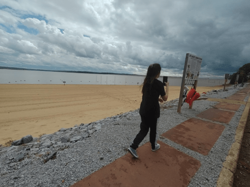 A person walks along a sandy beach with a cloudy sky, passing a sign and lifebuoys on the path.