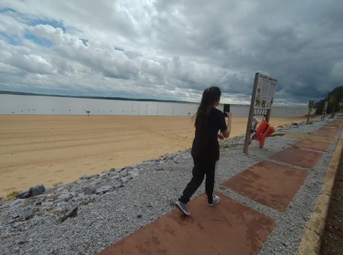 A person walks along a sandy beach with a cloudy sky, passing a sign and lifebuoys on the path.