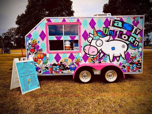 Colorful food truck with a unicorn design, serving ice cream, parked on grass with a menu sign nearby.