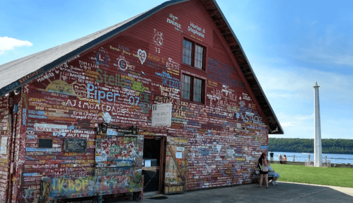 A colorful barn covered in painted signs and graffiti, with a lake and sky in the background. Two people sit nearby.