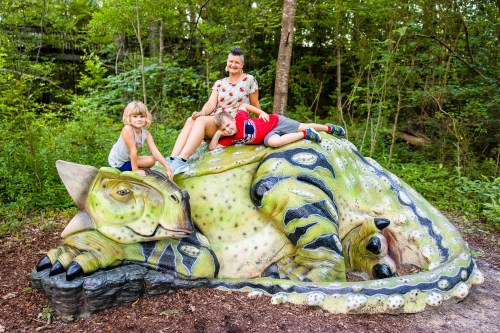 A woman and two children playfully pose on a large, colorful dinosaur sculpture in a lush, green forest.