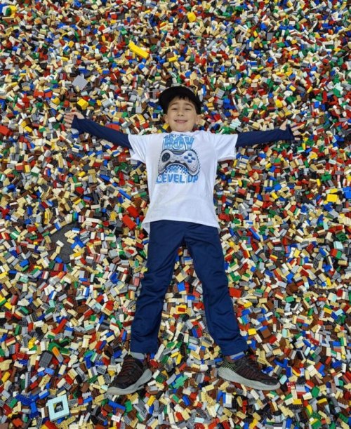 A child lies on a colorful pile of LEGO bricks, arms outstretched, smiling in a playful setting.