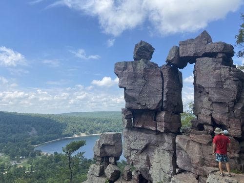 A person stands near a rock formation overlooking a lake and lush green hills under a blue sky with clouds.