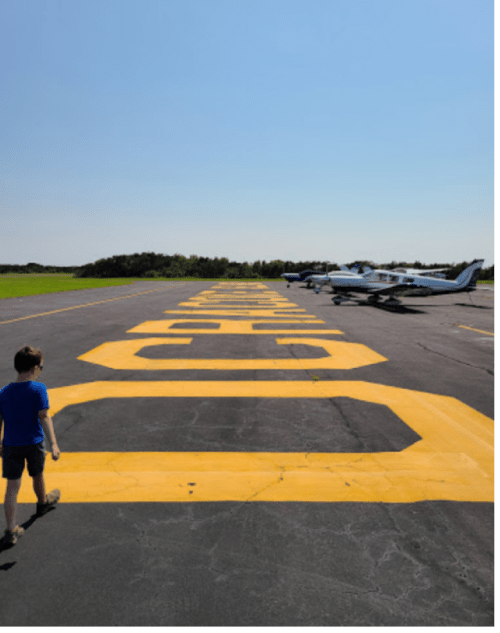 A child walks towards a runway marked with large yellow numbers, with small planes parked in the background under a clear sky.