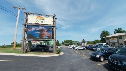 A zoo entrance with a large giraffe billboard and a parking lot filled with cars under a clear blue sky.