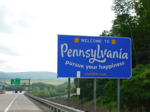 Blue welcome sign for Pennsylvania with the text "pursue your happiness" and a scenic background of hills and trees.
