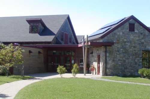 A modern stone and wood building with a sloped roof, solar panels, and landscaped greenery in front.