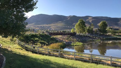 A serene landscape featuring a pond, green grass, trees, and mountains in the background under a clear blue sky.
