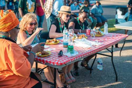 A group of four women enjoying a meal at a picnic table, with drinks and food on plates, in a lively outdoor setting.