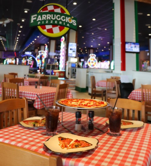 A cozy pizzeria with checkered tablecloths, featuring pizzas on a stand and drinks on the table. Bright arcade in the background.