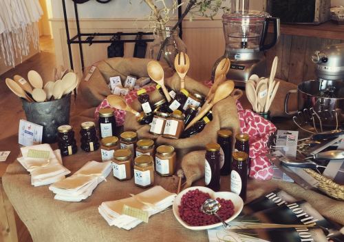 A rustic display of jars, wooden utensils, and kitchen tools on a table, featuring honey and other gourmet products.