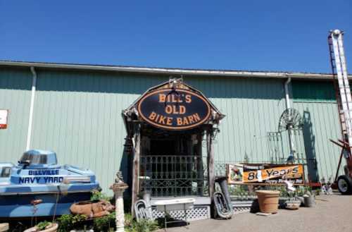 A rustic bike barn with a sign reading "Bill's Old Bike Barn," surrounded by plants and vintage items under a clear blue sky.