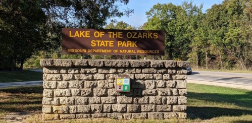 Sign for Lake of the Ozarks State Park, featuring stone base and surrounded by trees and grass.