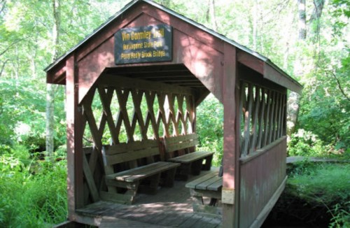 A wooden shelter with benches, surrounded by lush greenery on a trail. A sign reads "Van Beemley Trail."
