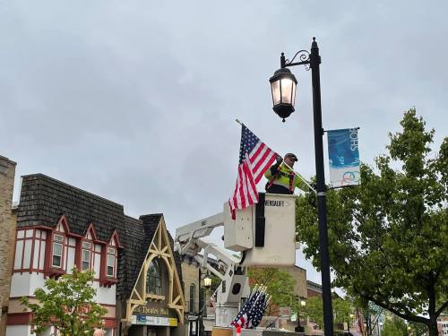 A worker in a lift hangs American flags on a lamp post in a town square, with buildings and cloudy skies in the background.