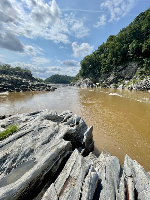 A serene river scene with rocky banks, lush green hills, and a partly cloudy sky reflecting on the water's surface.