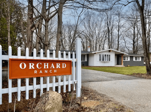 A modern house with a gray and white exterior, set behind a white picket fence with an "Orchard Ranch" sign.