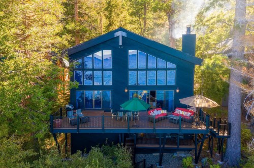 A modern black cabin surrounded by trees, featuring large windows and a deck with outdoor seating and umbrellas.