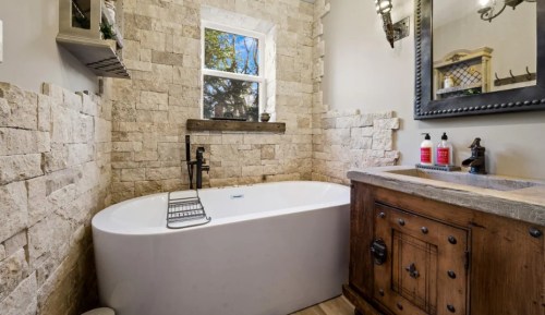 A modern bathroom featuring a freestanding tub, stone walls, and a wooden vanity with a sink. Natural light from a window.