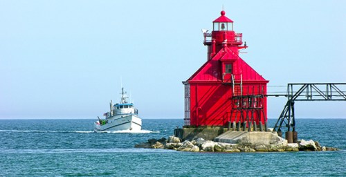 A red lighthouse stands on a rocky pier as a boat approaches in calm blue waters.