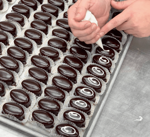 A person decorates chocolate-covered treats with white icing on a tray lined with paper cups.