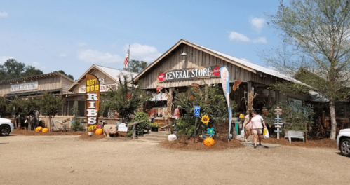 A rustic general store with signs for BBQ ribs, surrounded by trees and pumpkins, on a sunny day.