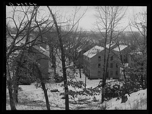 A snowy landscape featuring several buildings surrounded by bare trees, with a person walking in the foreground.