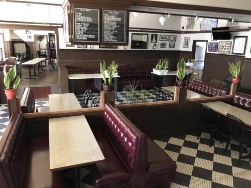 Interior of a diner featuring red booths, checkered floor, and potted plants, with a menu board on the wall.