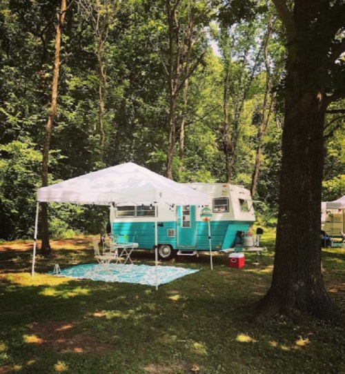 A vintage teal camper with a white canopy set up in a wooded area, surrounded by green trees and a picnic setup.