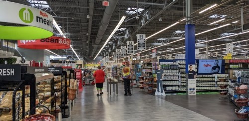 A spacious grocery store aisle with shoppers, shelves stocked with products, and bright overhead lighting.